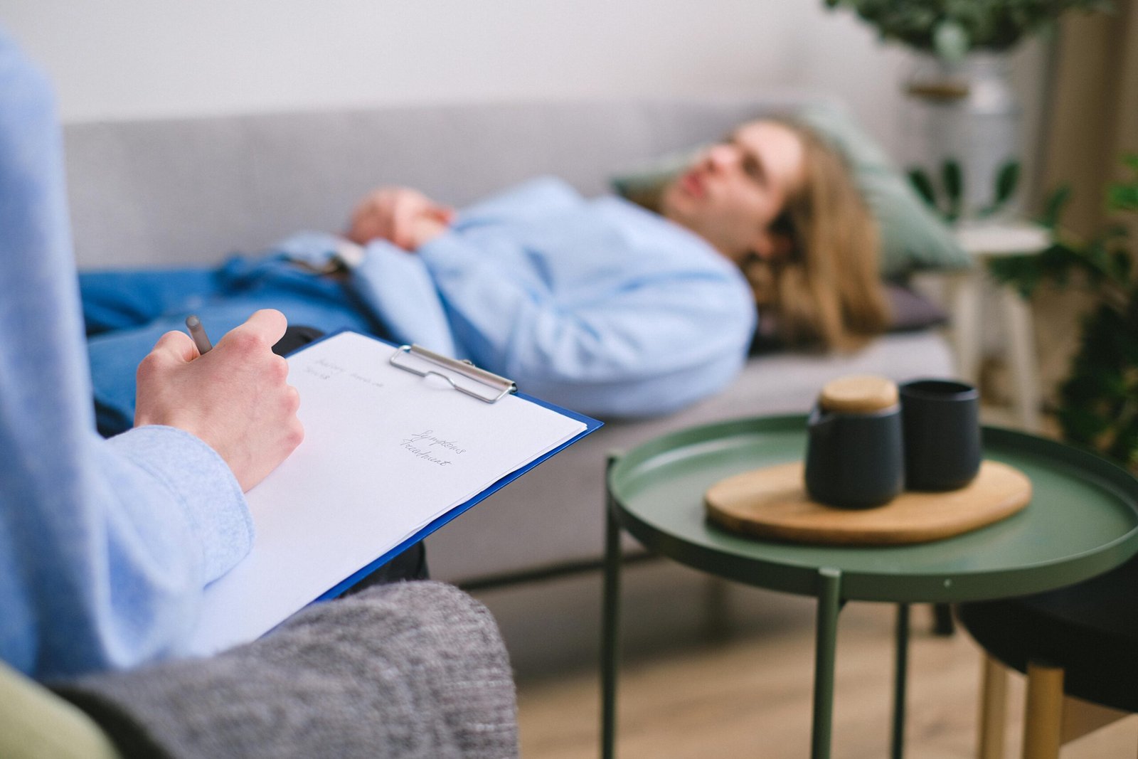 A therapist taking notes during a session with a patient lying on a sofa indoors.
